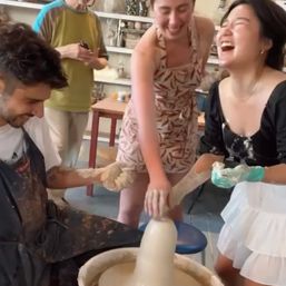 Group pottery class in a ceramics studio — three people laughing and shaping wet clay on a spinning pottery wheel, hands covered in clay with shelves of finished pieces in the background.