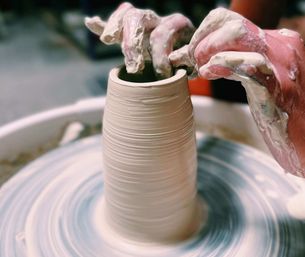 Close-up of hands shaping a wheel-thrown clay vase on a spinning potter's wheel in a pottery studio, wet clay and finger textures visible.