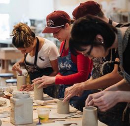 Hands-on pottery class in a bright ceramics studio — people in aprons shaping clay cups on a shared worktable.