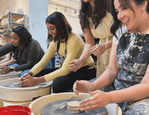 Group pottery wheel class — smiling women in clay-streaked aprons shaping bowls on electric wheels in a bright ceramics studio workshop.