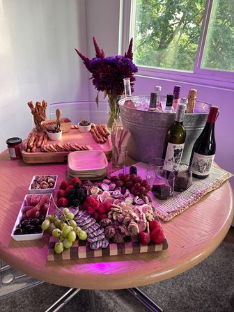 Party-ready charcuterie and wine spread on a round wooden table: grazing board with grapes, strawberries, blackberries, crackers, cookies, nuts and dips, pretzel sticks, bottles chilling in an ice bucket and a purple floral centerpiece by a sunlit window.