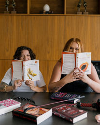 Two women seated at a wood-paneled table playfully peek over sex-positive books ('He Comes Next' and 'She Comes First'), surrounded by other sex-education books and adult toys in a cozy workshop-style setting.