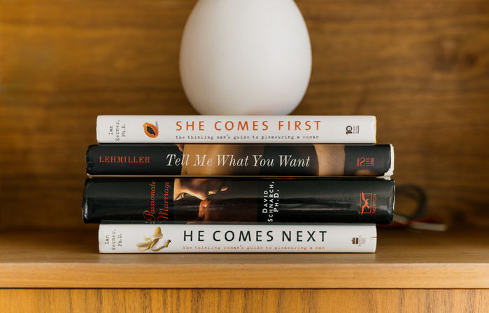 Stack of relationship and self-help books ('She Comes First', 'Tell Me What You Want', 'He Comes Next') on a wooden shelf topped by a white egg-shaped vase.