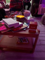 Cozy living room gathering around a low wooden coffee table with a glowing candle, bowls of snacks and popcorn, stacked books and magazines, and card game boxes under soft purple ambient lighting.