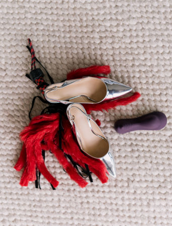 Playful scene of silver pointed-toe high heels, a red feather flogger and a purple silicone vibrator arranged on a chunky cream rug