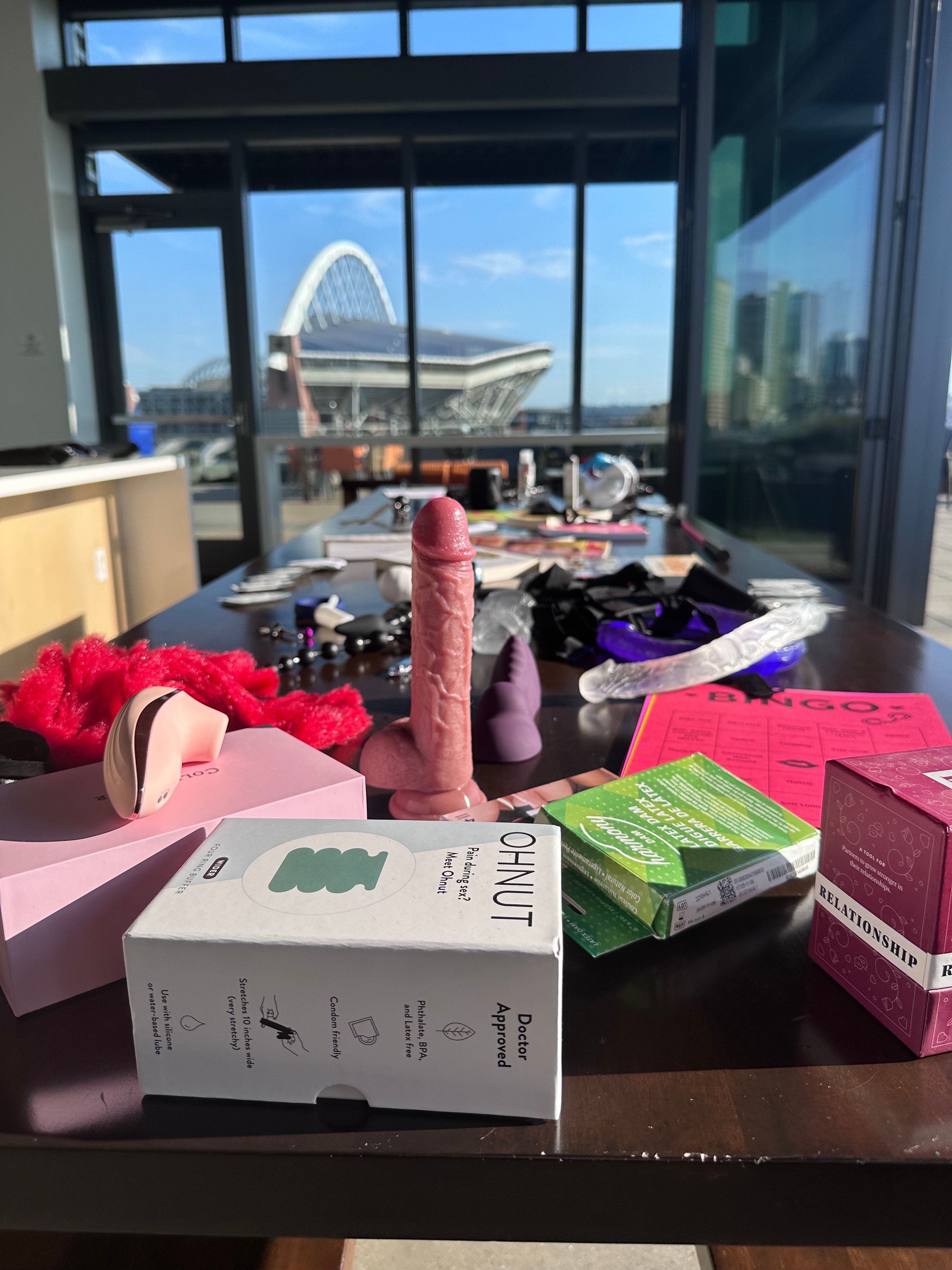 Sunlit condo table scattered with adult toys, boxes, and accessories — a pink dildo standing upright in the foreground, with floor-to-ceiling windows revealing an arched stadium and city skyline beyond.