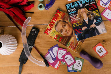 Flatlay of boudoir accessories on a wooden table: adult magazines, red feather tickler, leather paddle with chain, clear double-ended dildo, purple vibrator and playful intimacy game cards.