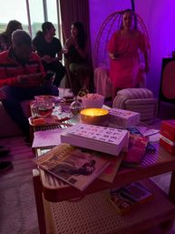 Cozy purple-lit living room gathering with friends chatting, a woman in a pink dress by a hanging chair, and a candlelit coffee table piled with books, snacks and a Skip‑Bo card game.