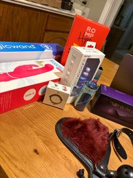 Assorted boxed adult toys and accessories arranged on a wooden table — pink vibrator packaging, small boxed gadgets, a canister, purple product box, and a red faux‑fur blindfold with straps and ball gag.