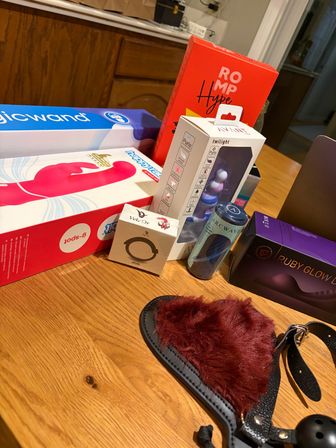 Assorted boxed adult toys and accessories arranged on a wooden table — pink vibrator packaging, small boxed gadgets, a canister, purple product box, and a red faux‑fur blindfold with straps and ball gag.