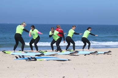 Beach surf lesson: group of five students in neon green rash guards practicing standing on surfboards on the sandy shore with an instructor and blue ocean waves in the background.