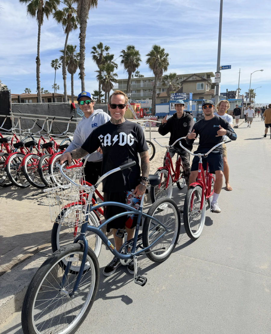 Group of five people on cruiser bicycles along a sunny Southern California beach boardwalk with palm trees, red rental bikes lined up and low-rise beachfront buildings in the background.