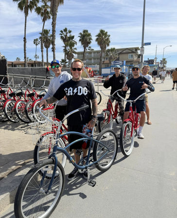 Group of five people on cruiser bicycles along a sunny Southern California beach boardwalk with palm trees, red rental bikes lined up and low-rise beachfront buildings in the background.