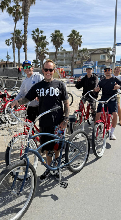 Group of five people on cruiser bicycles along a sunny Southern California beach boardwalk with palm trees, red rental bikes lined up and low-rise beachfront buildings in the background.