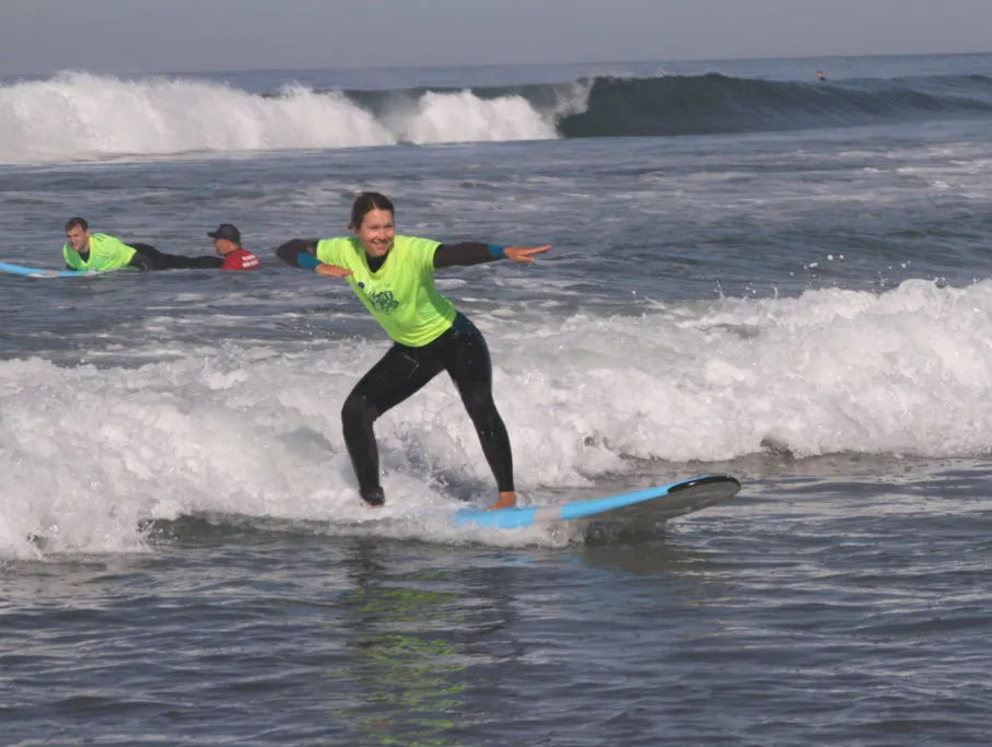 Smiling surfer in neon green shirt and black wetsuit balances on a blue surfboard riding a small shore break, other surfers and rolling ocean waves behind.