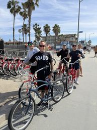 Smiling group of people with cruiser bikes on a sunny, palm‑lined oceanfront boardwalk with red rental bikes and beachfront buildings in the background.