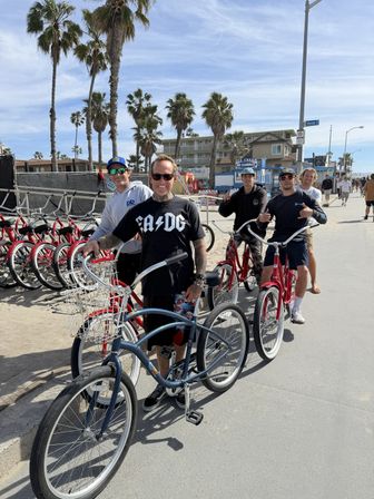 Smiling group of people with cruiser bikes on a sunny, palm‑lined oceanfront boardwalk with red rental bikes and beachfront buildings in the background.