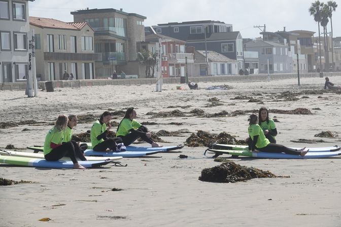 Group of people in bright green shirts sitting on surfboards on a sandy beach strewn with seaweed, with oceanfront houses and palm trees in the background — surf lesson vibes.