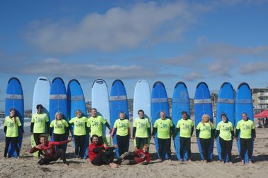 Cheerful surf school group in neon shirts lined up on a sunny sandy beach in front of tall blue surfboards with ocean and coastal buildings in the background