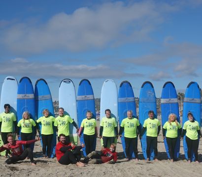 Cheerful surf school group in neon shirts lined up on a sunny sandy beach in front of tall blue surfboards with ocean and coastal buildings in the background