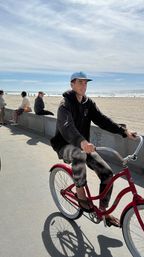 Person riding a red cruiser bicycle along a sunny oceanfront boardwalk with sandy beach and people on a concrete seawall