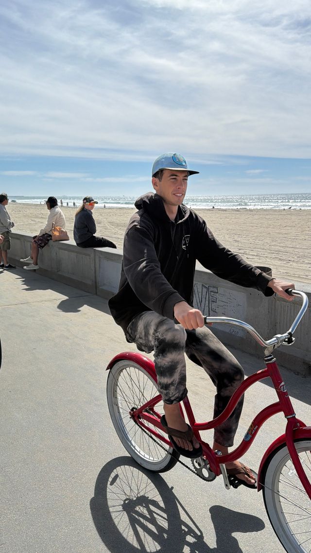 Person riding a red cruiser bicycle along a sunny oceanfront boardwalk with sandy beach and people on a concrete seawall