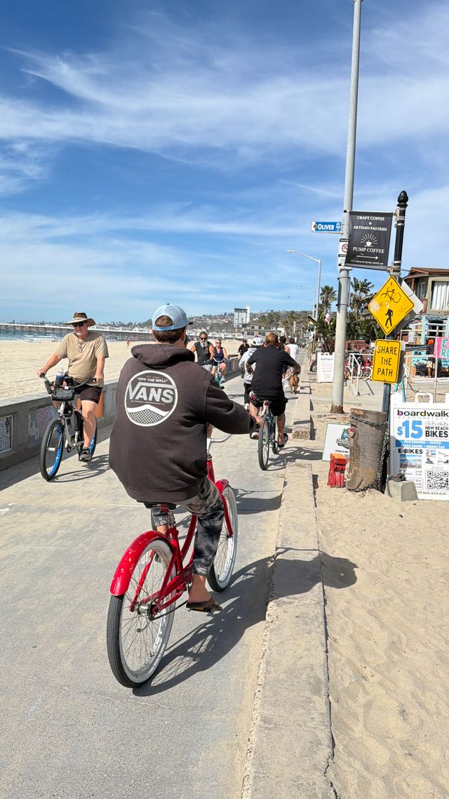 Sunny oceanfront boardwalk bike path with multiple cyclists — a rider in a black hoodie on a red cruiser, sandy beach and waves to the left, blue sky overhead, and beachside shops and signage lining the route.