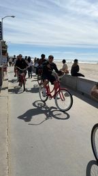Group riding red cruiser bikes along a sunny oceanfront boardwalk with sandy beach, sparkling waves and pedestrians in the background