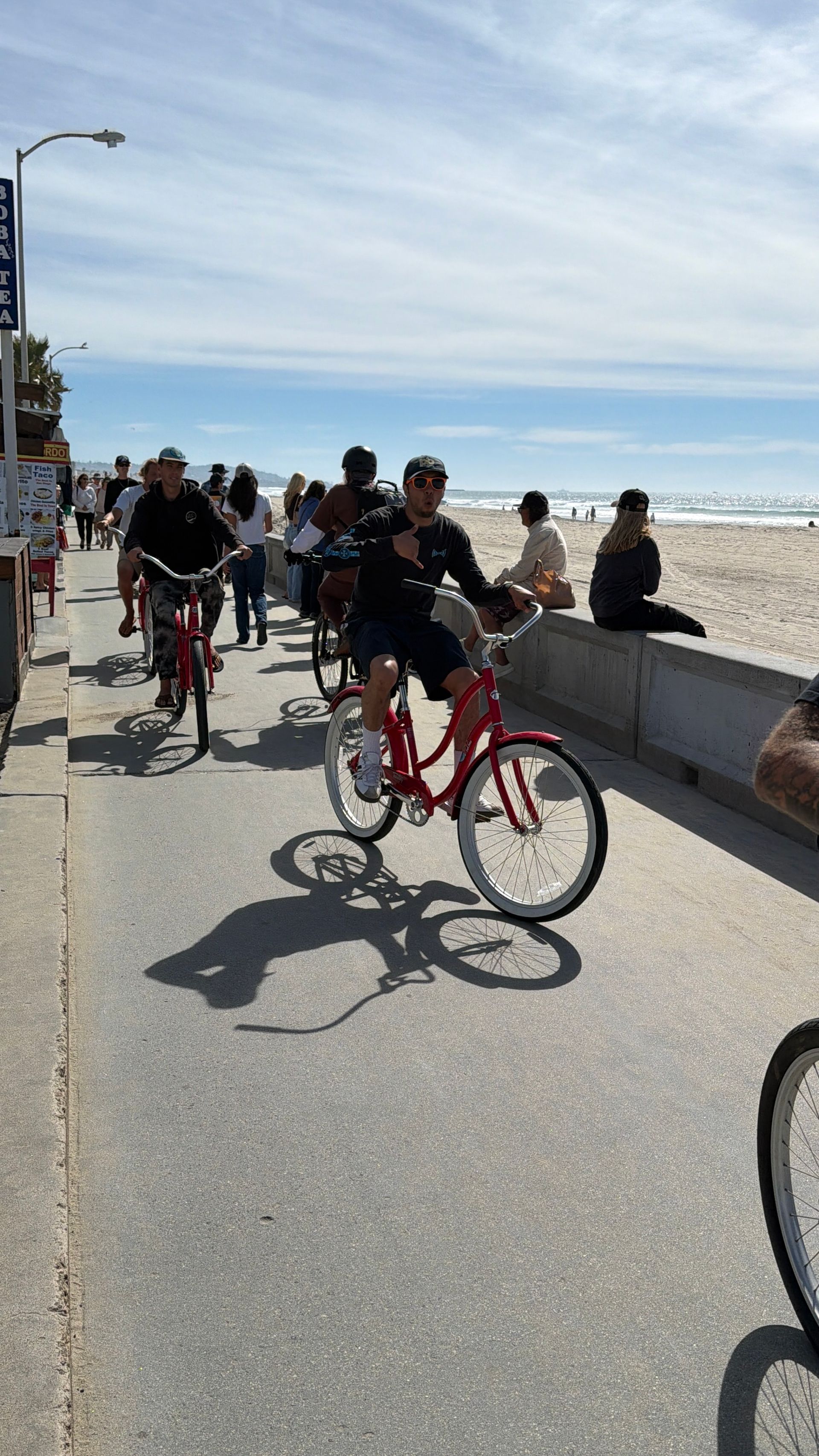 Group riding red cruiser bikes along a sunny oceanfront boardwalk with sandy beach, sparkling waves and pedestrians in the background