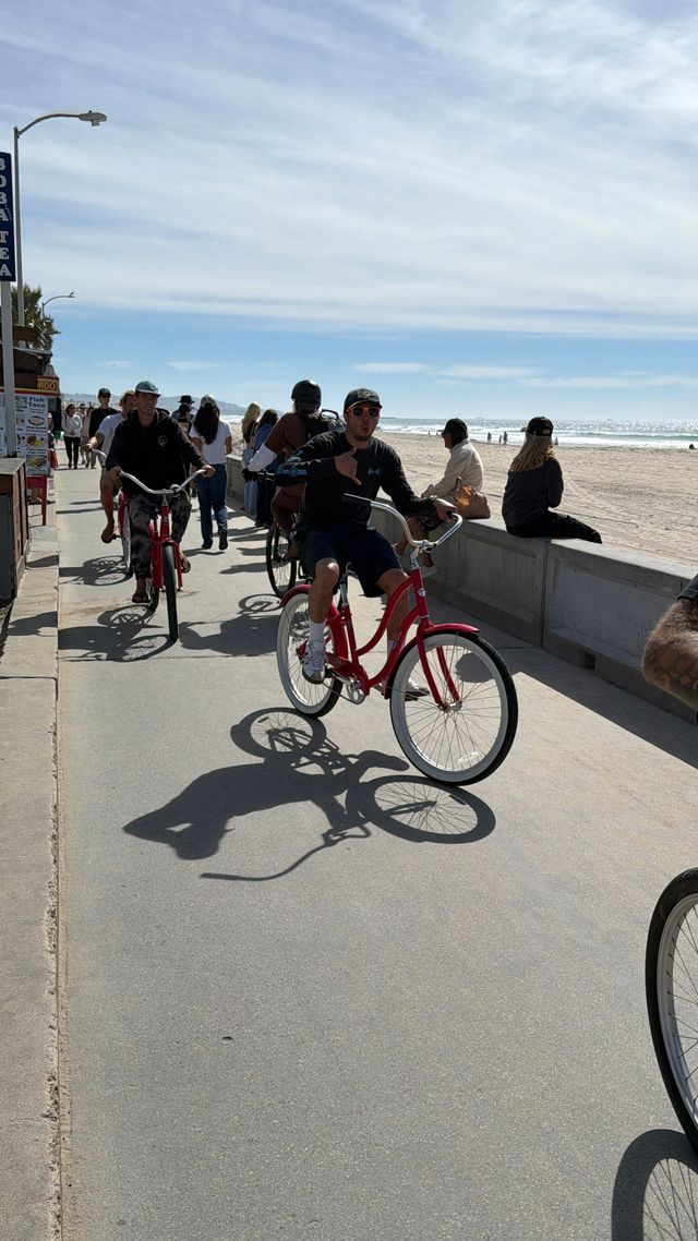 Group riding red cruiser bikes along a sunny oceanfront boardwalk with sandy beach, sparkling waves and pedestrians in the background