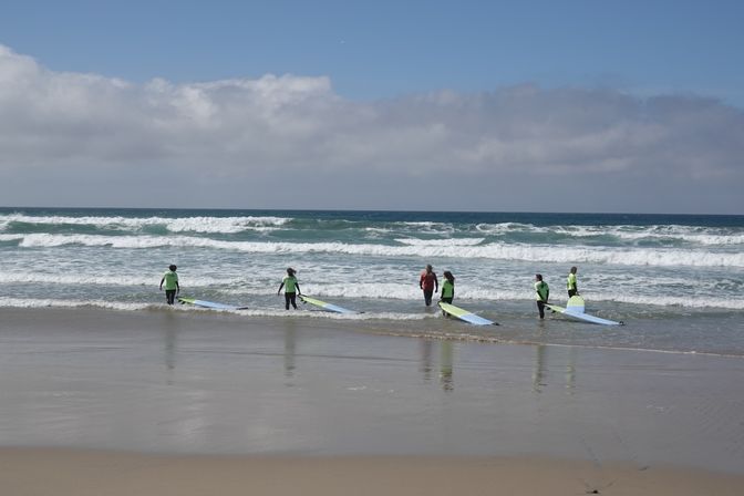 Group surf lesson with six people in wetsuits and bright rash guards carrying longboards into rolling ocean waves on a sandy coastal beach under a partly cloudy sky.