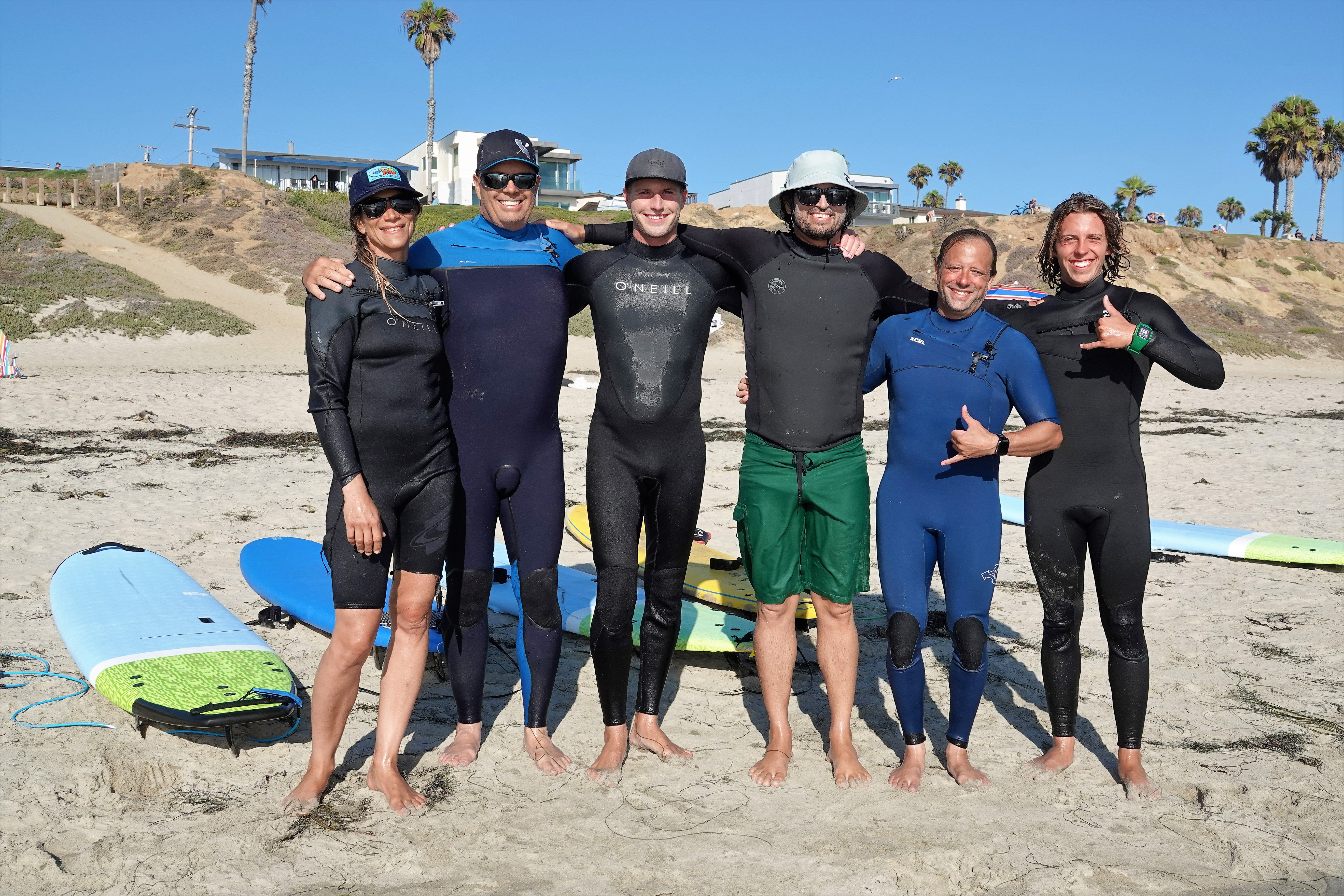 Six smiling surfers in wetsuits stand barefoot on a sunny California beach with colorful surfboards on the sand, palm trees and bluffside homes on the bluff behind them.
