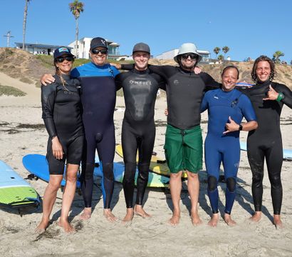 Six smiling surfers in wetsuits stand barefoot on a sunny California beach with colorful surfboards on the sand, palm trees and bluffside homes on the bluff behind them.