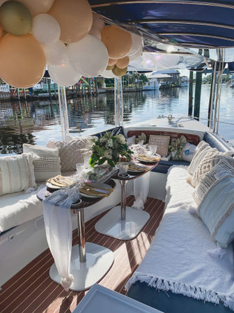 Cozy decorated boat interior with cream, white and gold balloons overhead, twin tables set with gold-accent plates, glassware and a floral centerpiece, textured pillows and throws, sunlight glinting on the marina water outside.