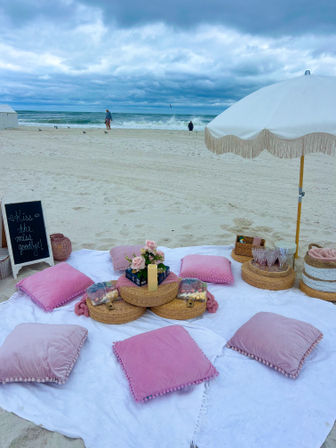 Cozy seaside picnic on a sandy beach with pink pillows, wicker trays, flowers and a fringed white umbrella, ocean waves and cloudy blue-gray sky in the background.