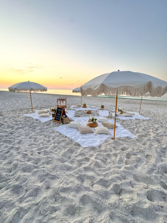 Boho beach picnic at sunset on a white-sand shore with fringed umbrellas, low pillows and blankets arranged near the calm ocean