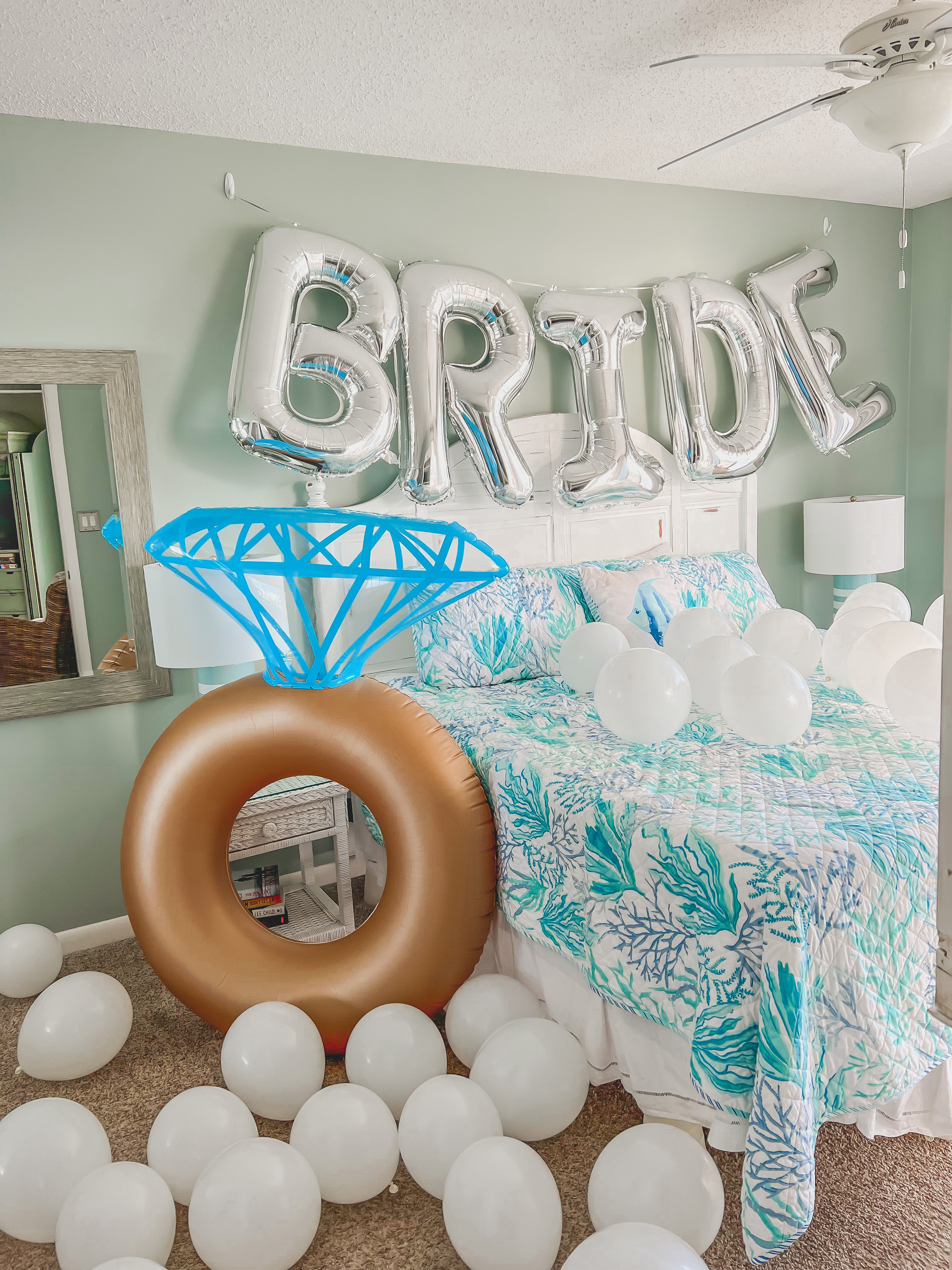 Coastal-style bedroom decked out for a bridal celebration with silver foil "BRIDE" balloons above a teal coral-print bed, scattered white balloons and a large inflatable gold engagement-ring prop