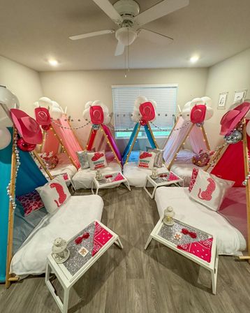 Indoor kids' slumber party setup in a bright room with six colorful teepee beds, pink cowgirl hats, white balloons and string lights, glittery pillows and small white tray tables with lanterns and pink bandanas on wood-look flooring