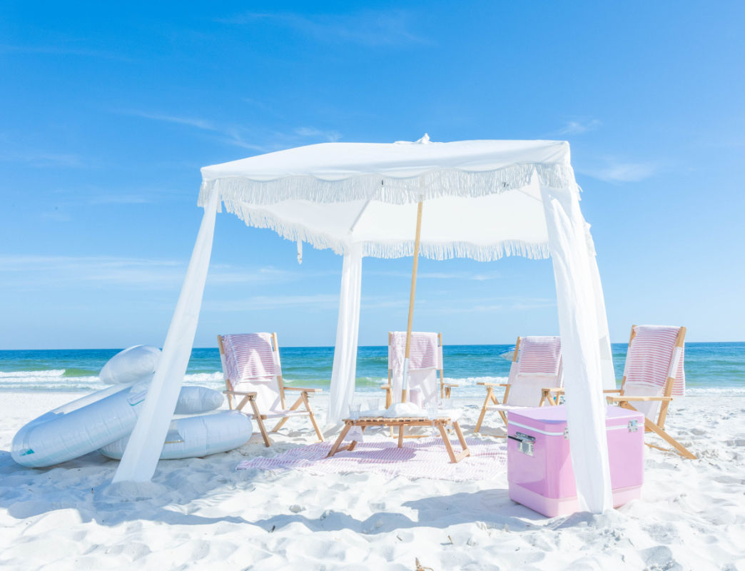 Vacation-ready white-fringed beach canopy with wooden lounge chairs, pink cooler and inflatable floaties on a sunny white-sand beach with turquoise ocean and clear blue sky