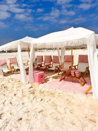 Chic white-sand beach scene with fringe-topped white canopies, wooden lounge chairs draped in red-and-white striped towels, matching striped mats and a pink cooler under a bright blue sky.