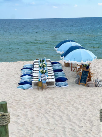 Oceanfront beach dining setup on white sand: low pallet table with blue-and-white place settings, patterned pillows, floral centerpieces and matching blue fringe umbrellas