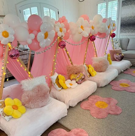 Indoor living-room slumber party setup with pink teepee tents draped in balloon daisy garlands, fairy lights and disco balls, plush fur and flower pillows on floor cushions and pink flower rugs.