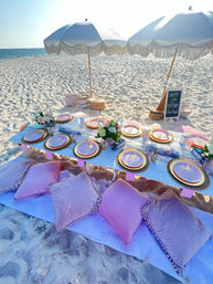 Sunny boho beach picnic on white sand — low white table set with pastel plates and gold chargers, pink velvet cushions, white fringed umbrellas, floral centerpieces and a birthday chalkboard by the ocean.