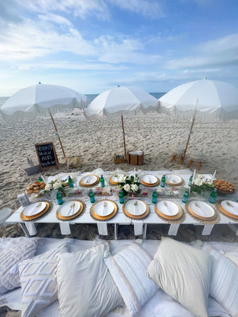 Beachfront boho picnic: low white table on sand set with gold-rim plates, floral centerpieces, bottled drinks, plush neutral pillows and white fringe umbrellas under a blue sky