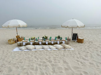Cozy boho beach picnic on a misty coastline — low wooden table set with plates, green glass bottles and floral centerpieces, surrounded by cushions and blankets under two white fringed umbrellas on soft sand with the ocean in the background.