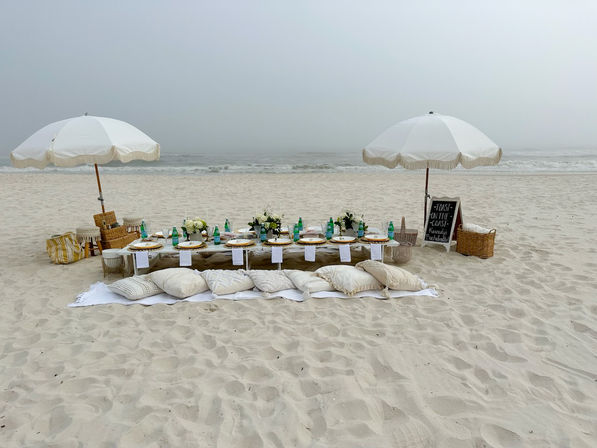 Cozy boho beach picnic on a misty coastline — low wooden table set with plates, green glass bottles and floral centerpieces, surrounded by cushions and blankets under two white fringed umbrellas on soft sand with the ocean in the background.