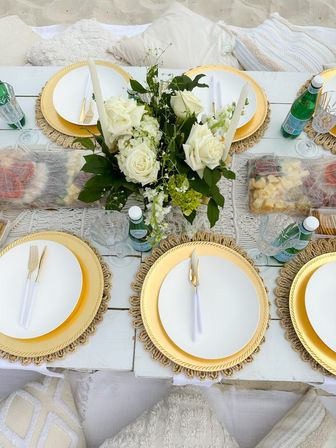 Beach picnic tablescape on sand with white rose centerpiece, gold charger plates, white dinnerware, gold flatware, crystal glasses and bottled sparkling water