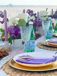Beachside table setting with purple taper candles, white roses and violet blooms, crystal glasses, sparkling water bottles, gold chargers, woven placemats and lavender napkins on white plates.