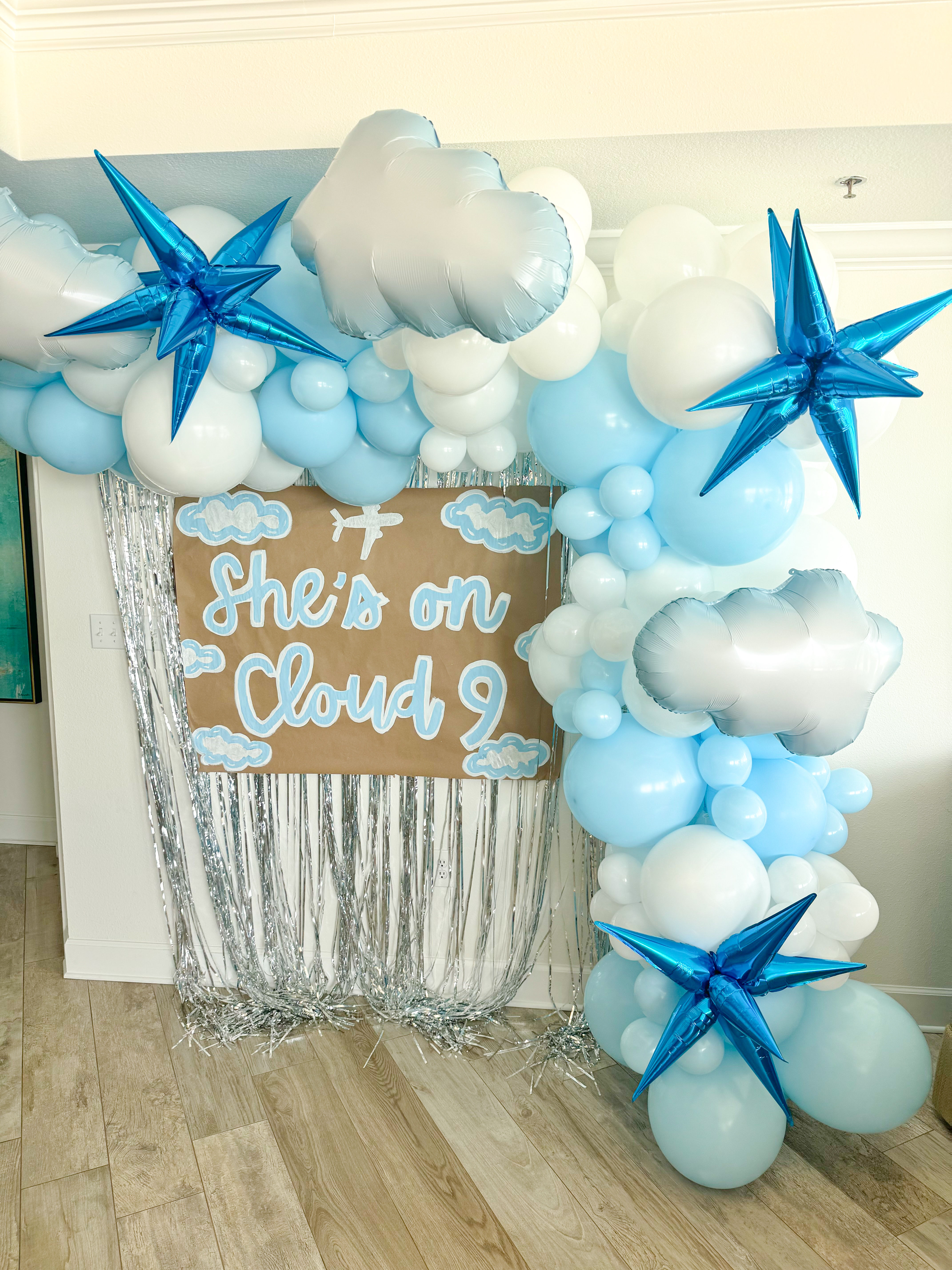 Indoor cloud-themed baby shower backdrop with blue and white balloon arch, silver cloud and star foil balloons, silver fringe curtain and kraft sign reading 'She's on Cloud 9'