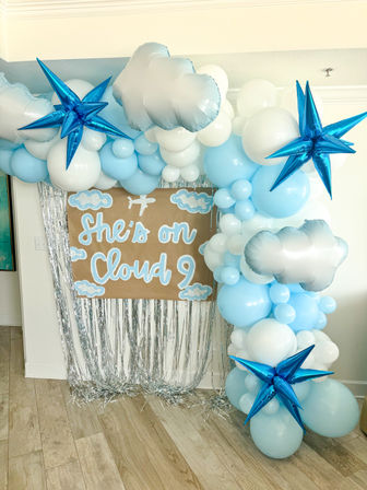 Indoor cloud-themed baby shower backdrop with blue and white balloon arch, silver cloud and star foil balloons, silver fringe curtain and kraft sign reading 'She's on Cloud 9'
